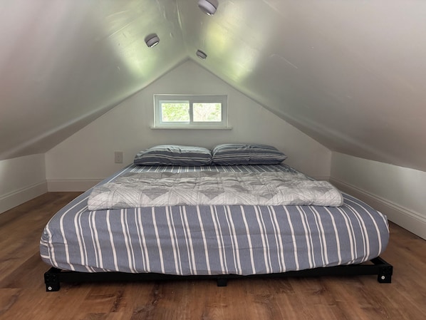 A bedroom with a sloped ceiling, a window, a bed with striped bedding, and a wooden floor.