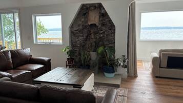 A living room with a stone fireplace, leather sofas, a coffee table, and large windows overlooking a body of water.