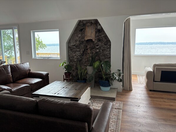 A living room with a stone fireplace, leather sofas, a coffee table, and large windows overlooking a body of water.