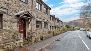 A stone building with a red door, a clear sky, and a road with cars. - Beddgelert Cottage Escape (Caernarfon)