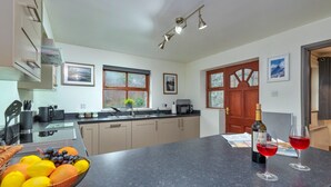 A modern kitchen with a central island, a dining area with a table set for two, and a view of the outdoors through the windows. - Beddgelert Cottage Escape (Caernarfon)