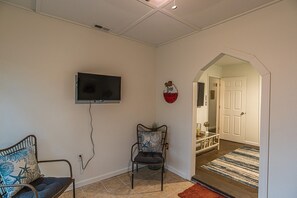 Ein Zimmer mit einem an der Wand befestigten Fernseher, einem Sessel mit Kissen und einer Tür zu einem weiteren Raum.