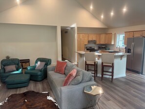 A modern living room with a grey sofa, teal armchairs, and a glass coffee table. A kitchen with wooden cabinets and stainless steel appliances is visible in the background. - Brand new, bright 4-bedroom house near ski hills! (Harbor Springs)