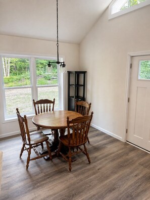 A dining area with a wooden table and chairs, a hanging light fixture, and a view of the outdoors through large windows. - Brand new, bright 4-bedroom house near ski hills! (Harbor Springs)