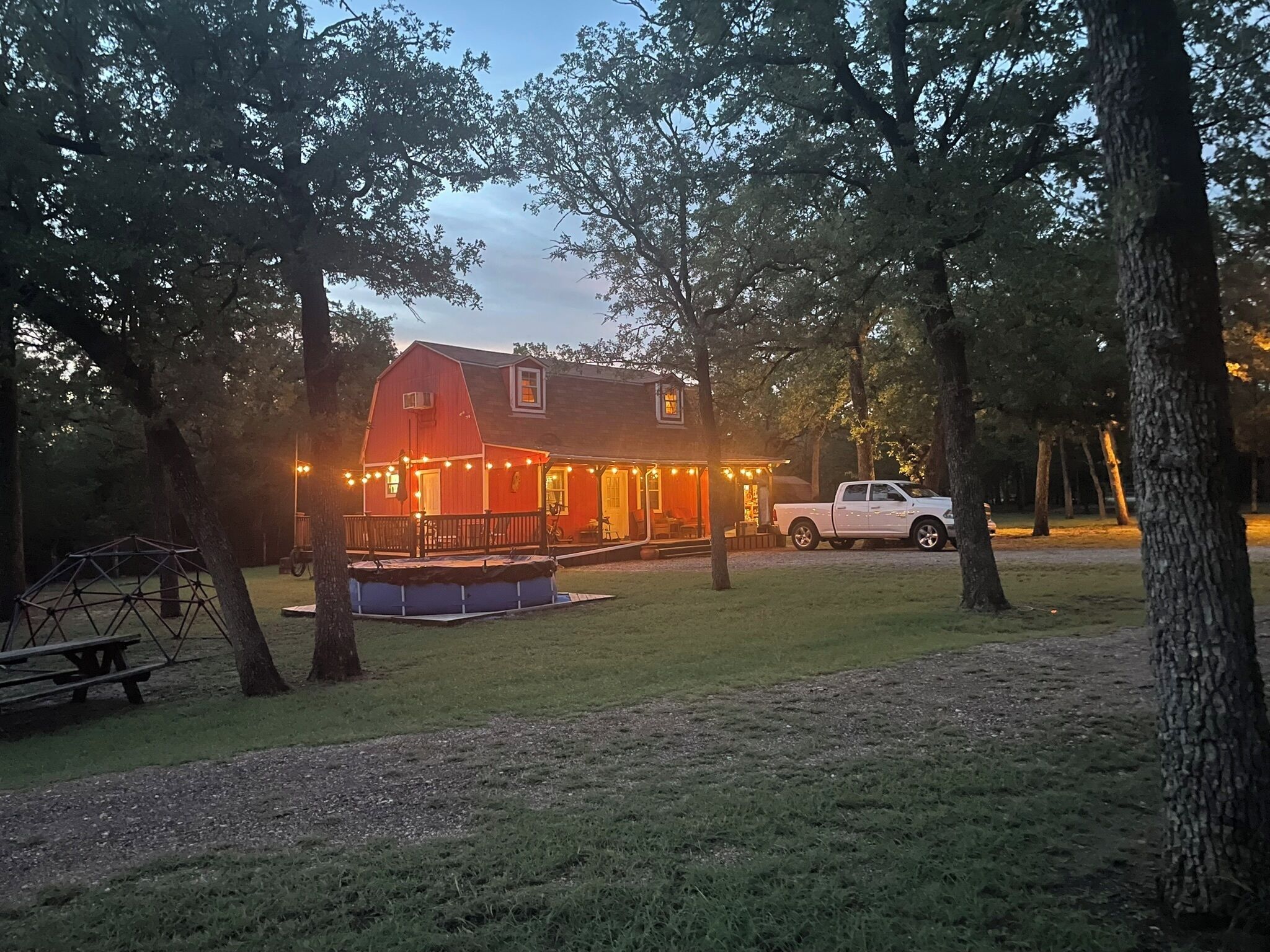 A red barn with a porch and string lights, surrounded by trees and a white pickup truck.