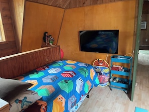 Ein Zimmer mit einem Bett, einem an der Wand befestigten Fernseher und einem blauen Rollwagen mit Gegenständen darauf.