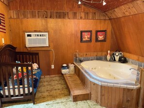 Ein Holzblockhaus-Interieur mit Whirlpool, Babybett mit Stofftieren und zwei gerahmten Bildern an der Wand.