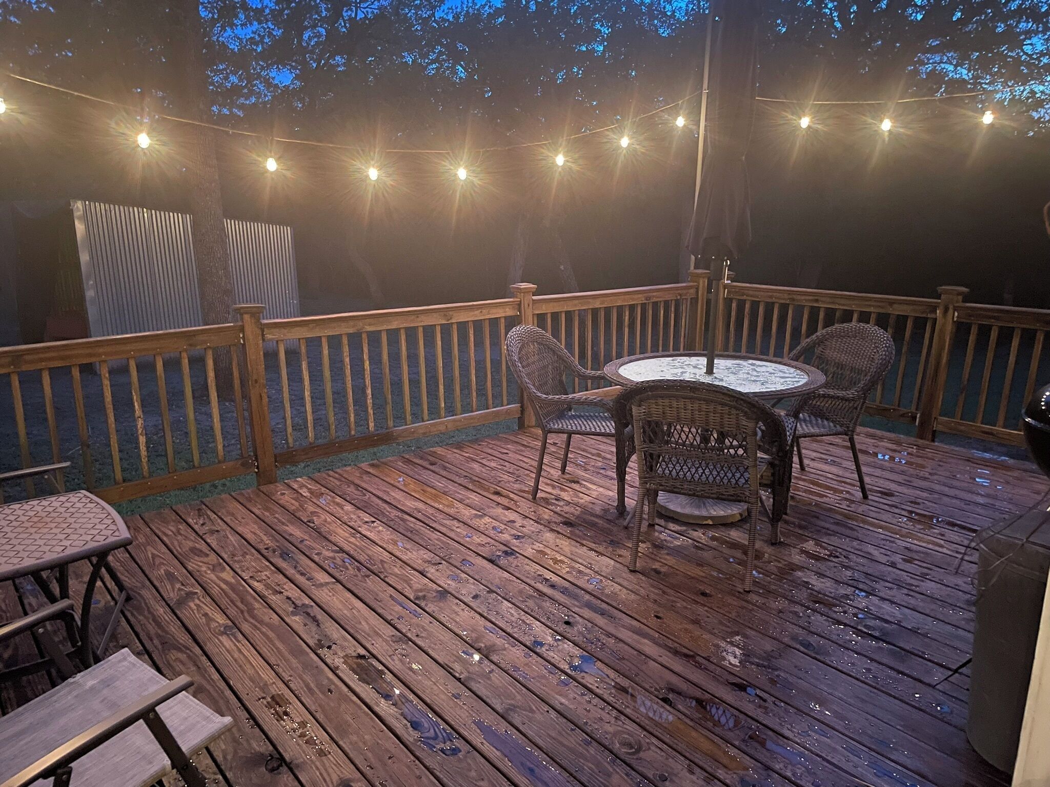 Una terraza de madera con una mesa redonda y sillas de mimbre, rodeada por una barandilla de madera y luces colgantes.