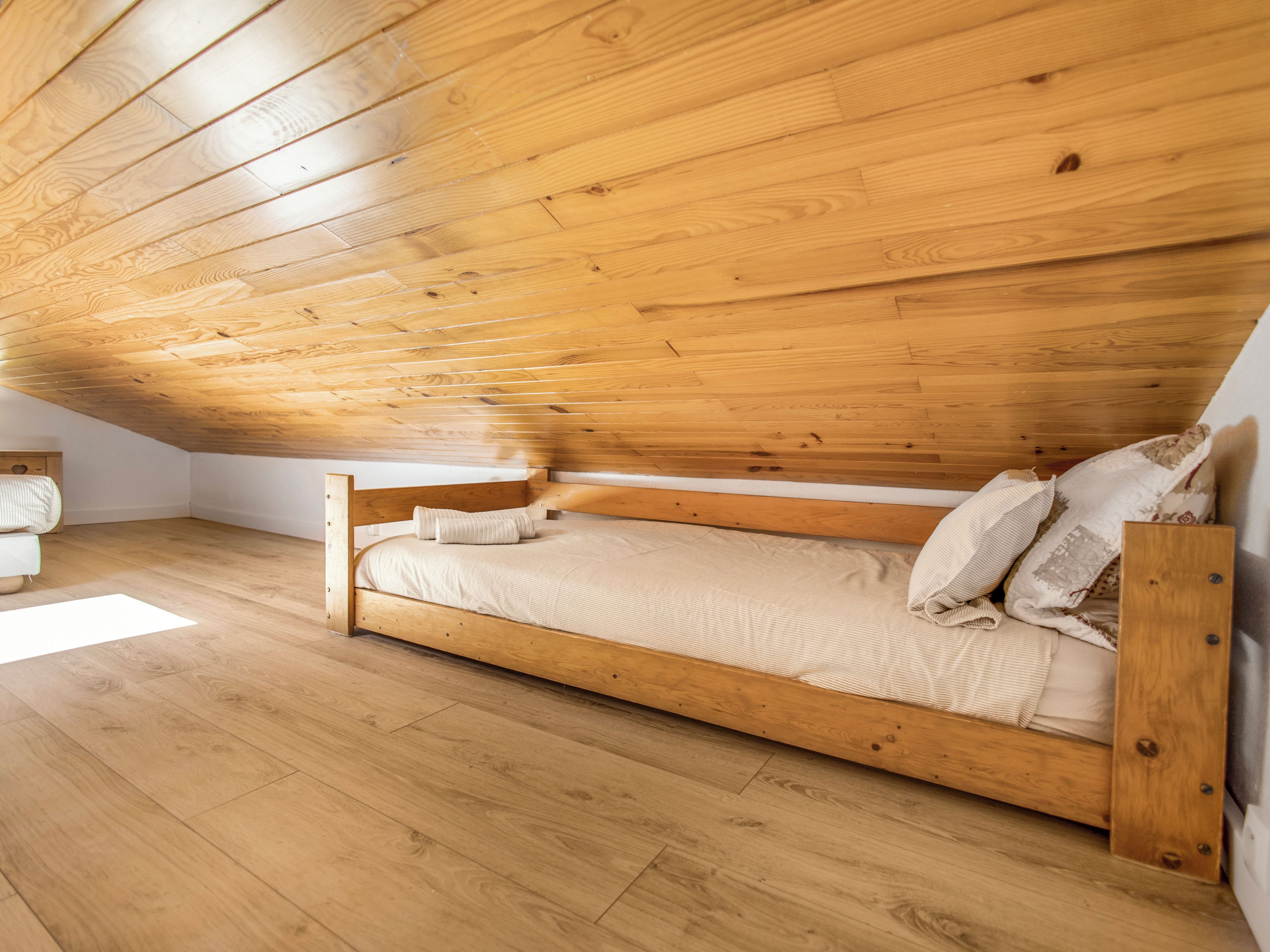 Camera con un letto singolo, soffitto e pavimento in legno, un letto semplice con materasso e cuscini, e un comodino con lampada.