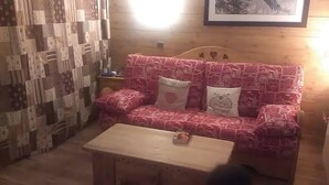 Ett mysigt rum med en väggpanel av trä, en mönstrad täckfilt, en röd soffa med dekorativa kuddar och ett litet träbord.