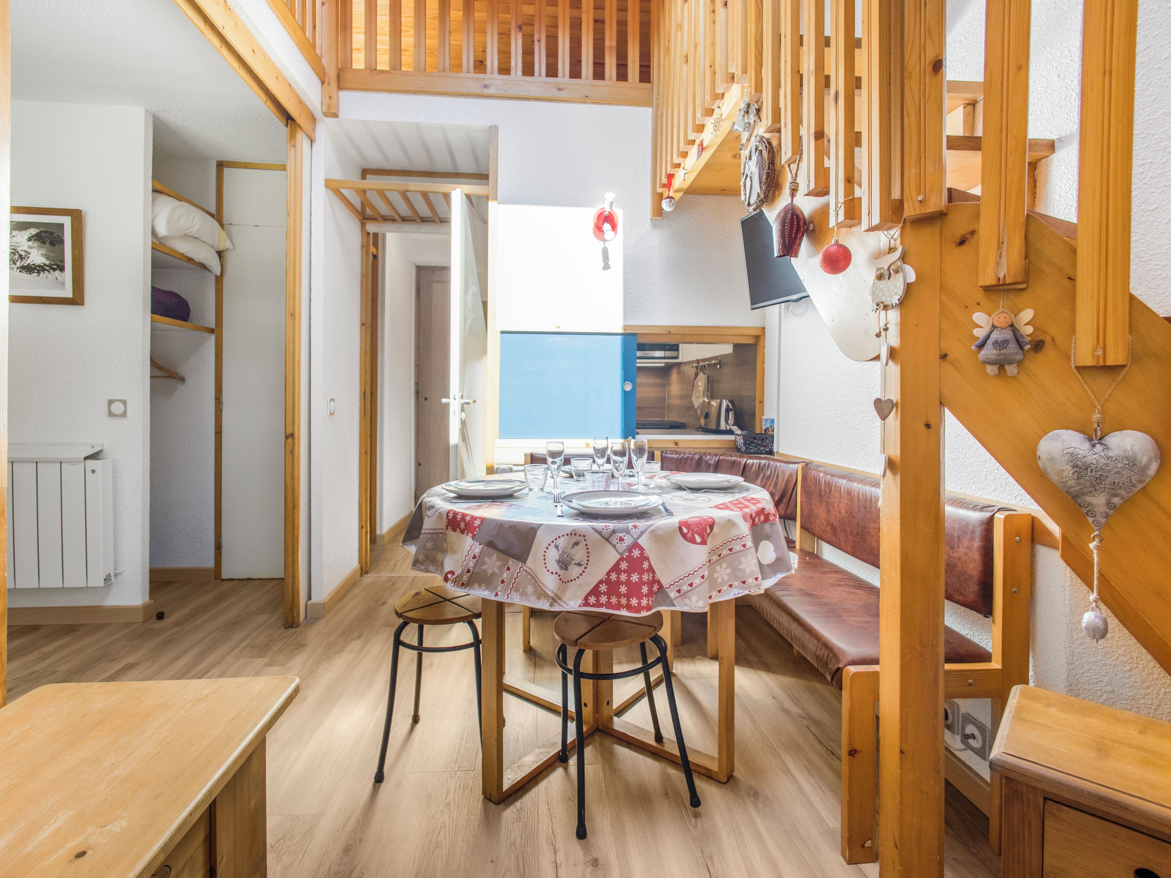 Un coin repas avec une table ronde dressée pour quatre personnes, un plancher en bois et une vue sur la cuisine grâce à une porte ouverte.