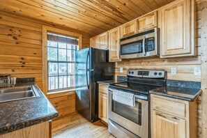 A compact kitchen with wooden walls, a stainless steel refrigerator, a microwave, an oven, and a granite countertop.