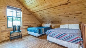 A room with wooden walls and a sloped ceiling, featuring two beds with different bedding and a small table with speakers. - Premium Cabin - Family Cabin Rental Sleeps 8-10 (Willis)