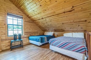 A room with wooden walls and a sloped ceiling, featuring two beds with different bedding and a small table with speakers. - Premium Cabin - Family Cabin Rental Sleeps 8-10 (Willis)