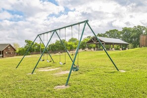 A swing set in a grassy area with a wooden structure in the background. - Premium Cabin - Family Cabin Rental Sleeps 8-10 (Willis)