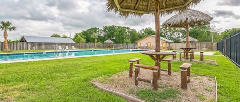 Un patio trasero con piscina, mesas de picnic y un quiosco de paja.