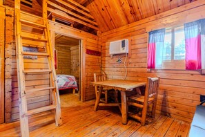 Interior de una cabaña de madera con una mesa y sillas de madera, una escalera, un equipo de aire acondicionado y una ventana con cortinas.
