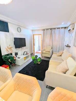 A living room with a sofa, armchairs, a TV, a plant, and a kitchen area with a sink and cabinets. - STYLE AND COMFORT IN THE HEART OF THE CITY (LOME)