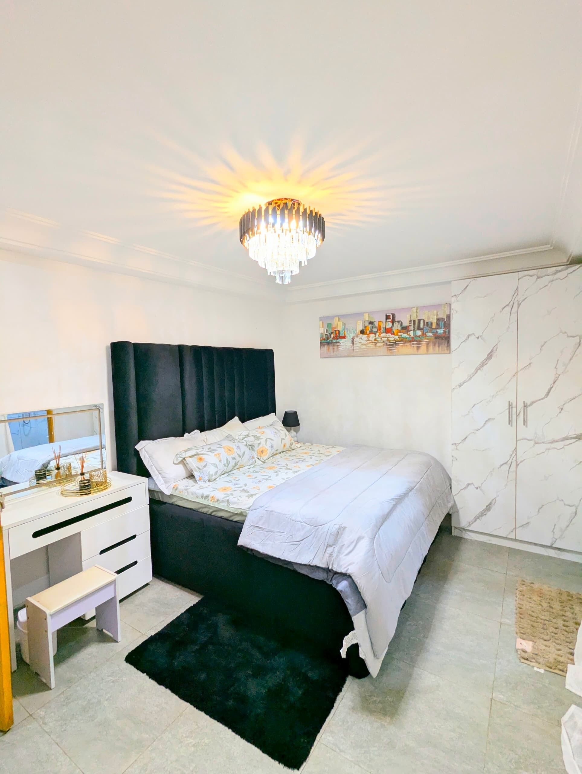 A bedroom with a bed, a desk, a chair, a wardrobe, a chandelier, and a cityscape painting.