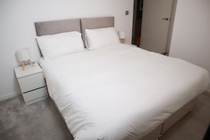 A neatly made bed with a headboard, a bedside table with a lamp, and a small drawer unit. - Luxury Modern Flat BHX NEC JLR Solihull Hospital with Free Secured Parking (Solihull)