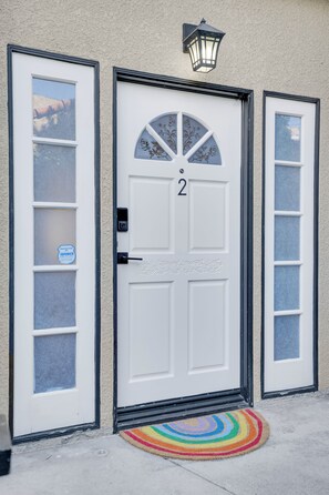 A white double door with a number '2' above it, a small window with a decorative design, and a colorful doormat on the ground. - Spacious 2BR/2.5BA Coastal Retreat- Parking, Walkable to Water, Near Highways (Los Angeles)