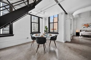 Ein modernes Interieur mit Essbereich, der ein Glastisch und schwarze Stühle, eine Treppe sowie ein Schlafzimmer mit Bett und Gemälde zeigt.