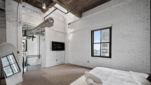 A room with white brick walls, a large window, a bed with white bedding, and a visible air duct. - Top-Floor Downtown Penthouse - Terrace - 2,500 Sq - Gym+Pool+Dedicated Workplace (Dallas)