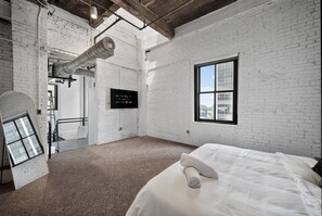 Ein Zimmer mit weißen Backsteinwänden, einem großen Fenster, einem Bett mit weißer Bettwäsche und einem sichtbaren Lüftungskanal.