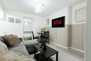 A modern living room with a sofa, a coffee table, and a dining area with chairs. A television is mounted on the wall displaying the Netflix logo. - Modern 3BR | Sleeps 6 | Free Parking | Central London (London)