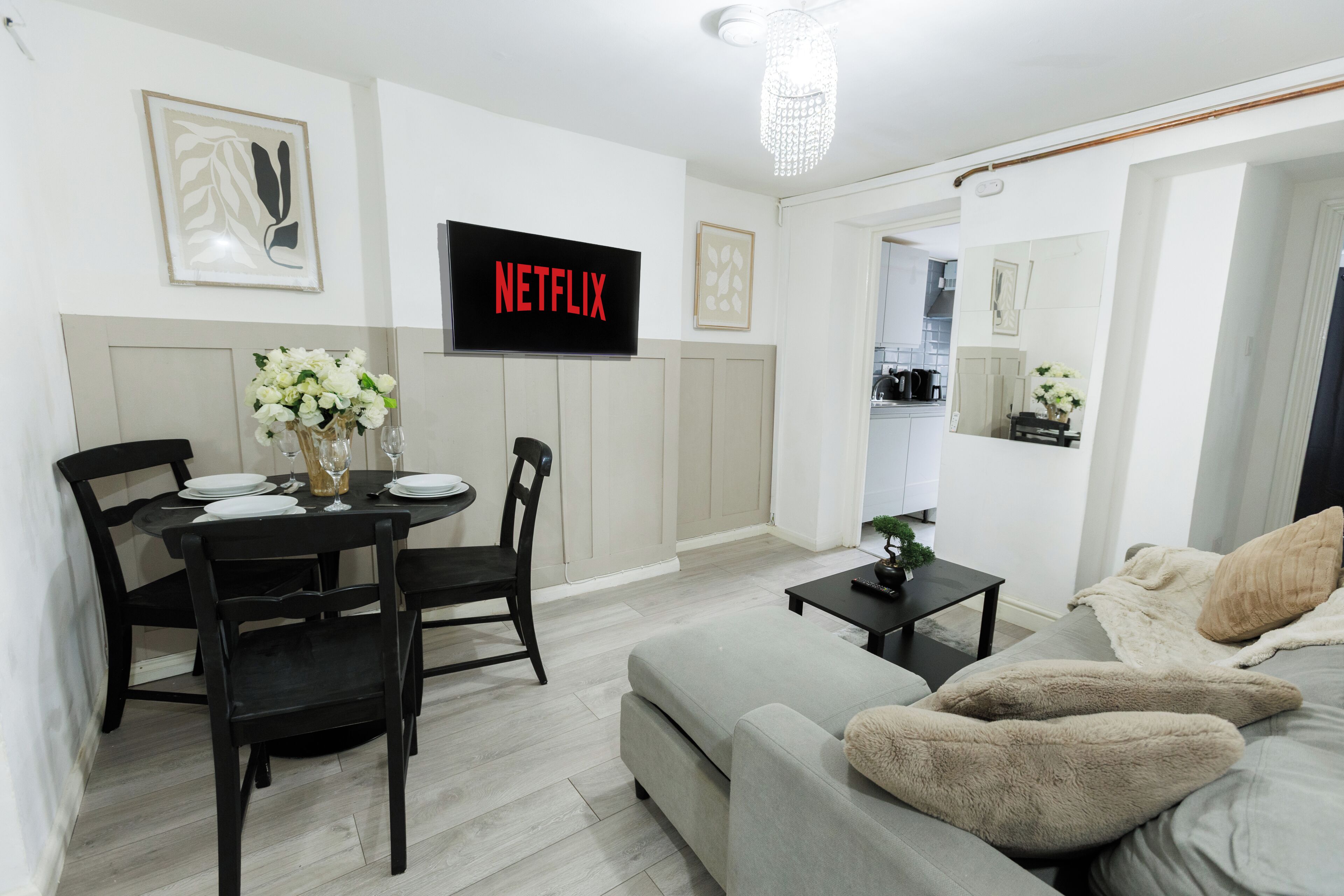 A modern living room with a grey sofa, a black dining table with chairs, a flat-screen TV mounted on the wall, and a small potted plant on a side table.