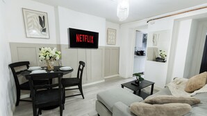 Uma sala de estar moderna com um sofá cinzento, uma mesa de jantar preta com cadeiras, uma TV de tela plana na parede e uma pequena planta em vaso sobre uma mesa lateral.