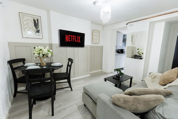 Ein modernes Wohnzimmer mit grauem Sofa, einem schwarzen Esstisch mit Stühlen, einem flachbildfernseher an der Wand und einem kleinen Blumentopf auf einem Beistelltisch.