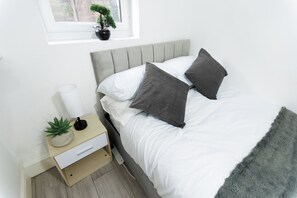 Una cama bien hecha con ropa de cama blanca y gris, una mesita de noche con una lámpara, una planta en maceta y una regleta de enchufes.