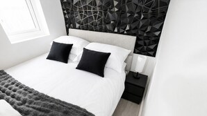 Quarto moderno com cabeceira estampada em padrão geométrico, cama com roupa de cama branca, almofadas pretas e manta cinza.