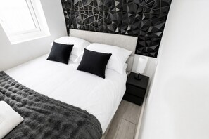 Ein modernes Schlafzimmer mit einem Kopfteil im geometrischen Muster, einem Bett mit weißer Bettwäsche, schwarzen Kissen und einer grauen Tagesdecke.