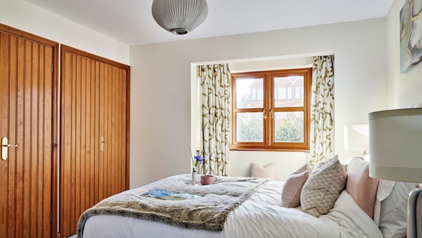 Ein Schlafzimmer mit großem Holzschrank, einem Bett mit weißer Bettdecke und mehreren Kissen, einem Fenster mit Vorhängen und einer Hängeleuchte.
