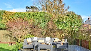 Eine Holzterrasse mit einem grauen Ecksofa, einem Couchtisch und einem kleinen Hund.