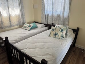 Ein Doppelbett mit weißer Bettwäsche, zwei Zierkissen, einem Holz-Kopfteil und einem Fenster mit gemusterten Vorhängen.