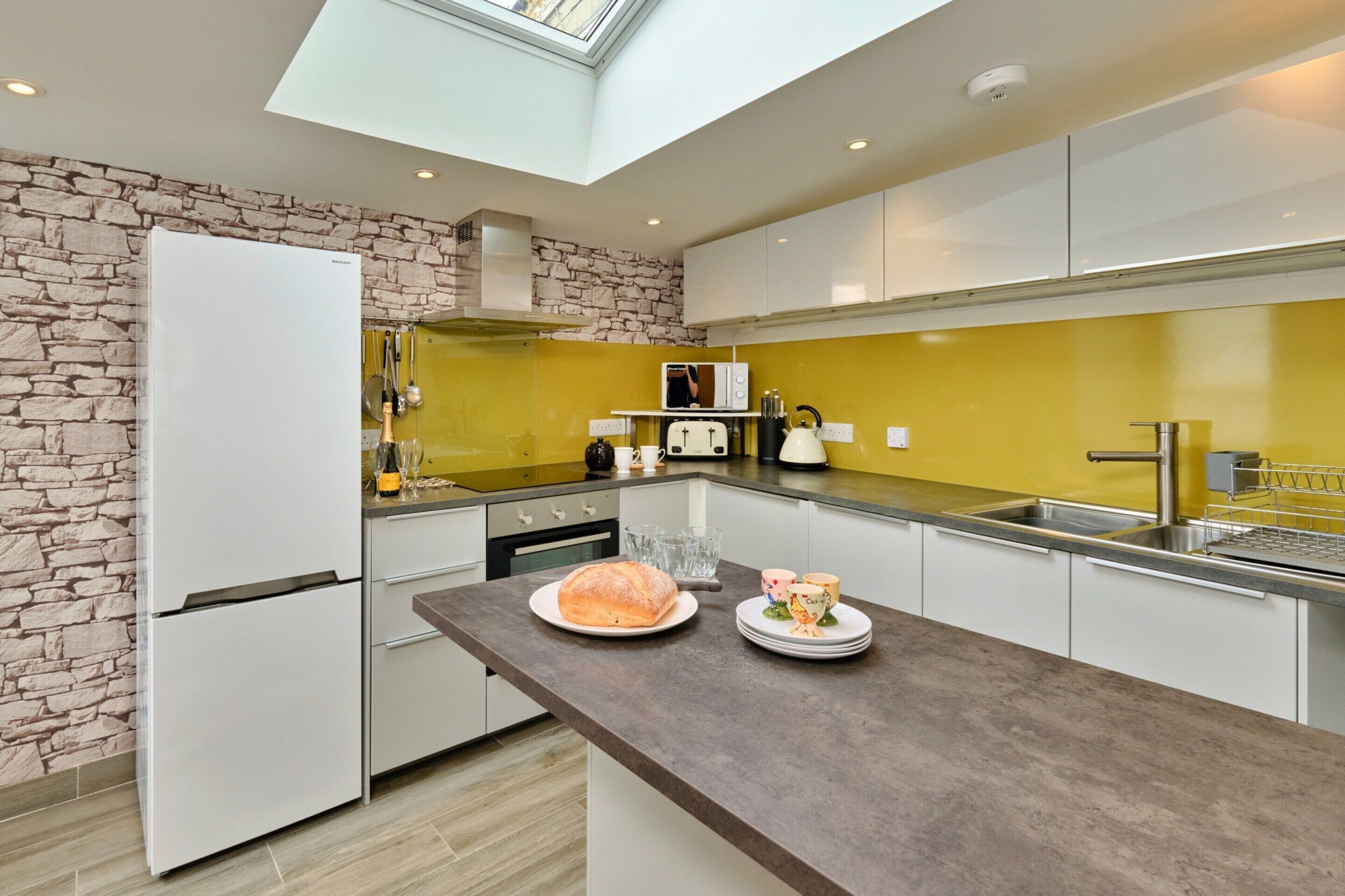 A modern kitchen with a stone wall, a skylight, and a central island with a loaf of bread and plates.