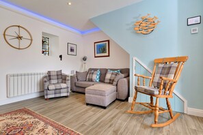A living room with a sectional sofa, a wooden rocking chair, a wall clock, and a radiator. - Grand Devon Country House for 15 (Penzance)