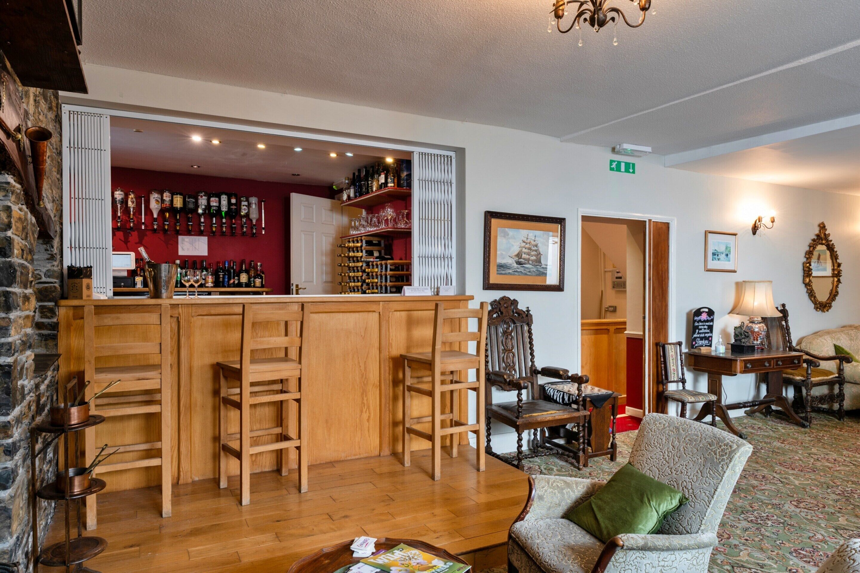 Una zona de bar con taburetes de madera, una barra con botellas y una pared roja. También hay sillas y una mesa con una lámpara.