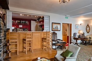 Una zona de bar con taburetes de madera, una barra con botellas y una pared roja. También hay sillas y una mesa con una lámpara.