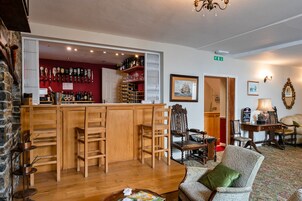 Una zona de bar con taburetes de madera, una barra con botellas y una pared roja. También hay sillas y una mesa con una lámpara.