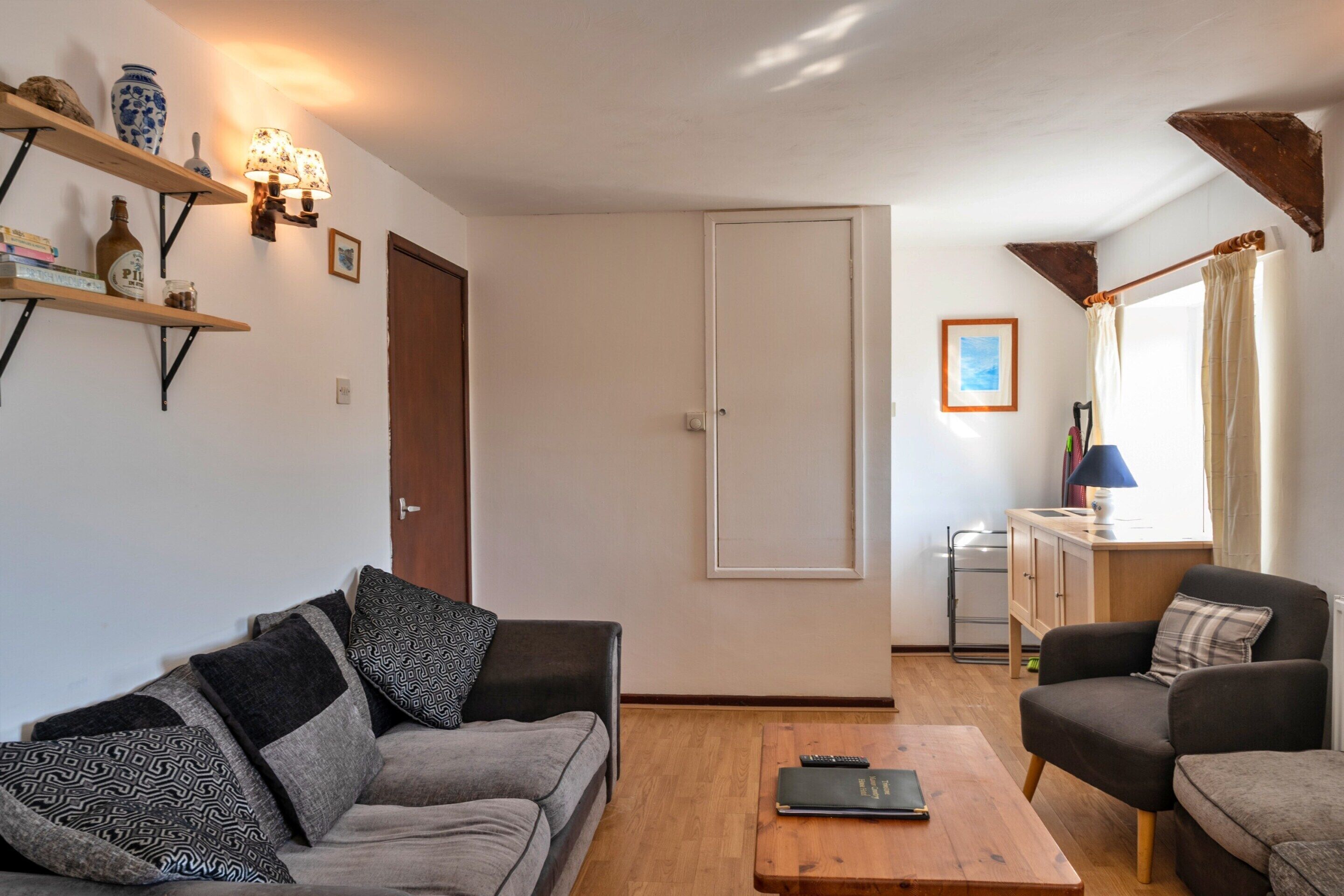 Una sala de estar acogedora con un sofá, una mesita de centro y una mesita auxiliar con una lámpara. Hay una puerta que da a otra habitación y una ventana con cortinas.