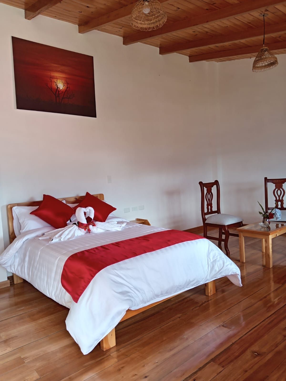 Una cama bien hecha con ropa de cama roja y blanca, suelo de madera, una silla de madera y una mesa con un florero.