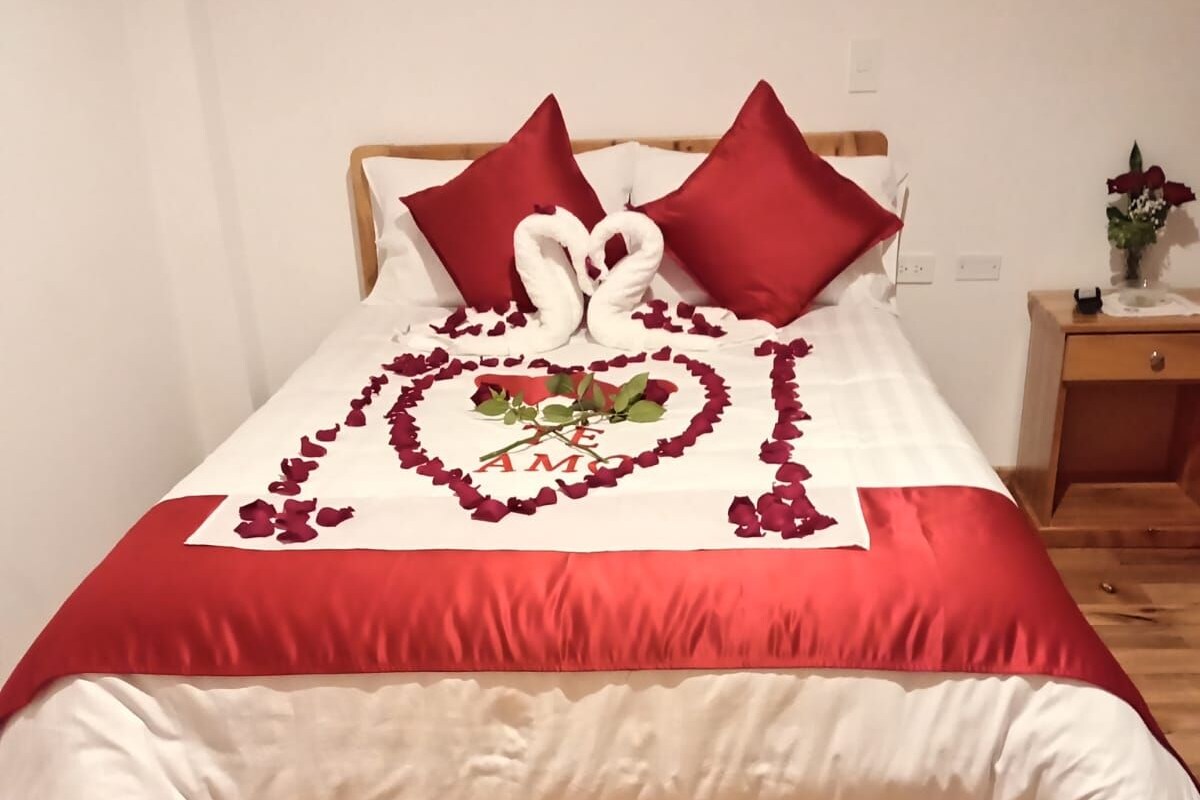 Een bed met een hartvormige bloemblaadjesdecoratie van rozen en een enkele roos, een houten hoofdbord en een nachtkastje met een bloevas.