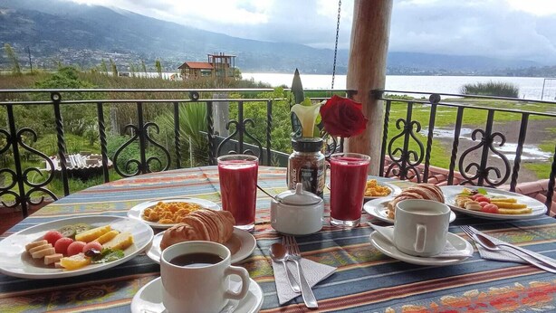 A table set with breakfast items, including croissants, fruit, and beverages, overlooking a scenic landscape. - Brisas Del Lago San Pablo (Otavalo)