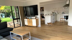 A modern living room with a sofa, a coffee table, and a TV. There is a kitchen area with cabinets and a stove. A cat is visible in the room. - Relaxing haven for Families - minutes walk to the beach, shops & dining (Tauranga)
