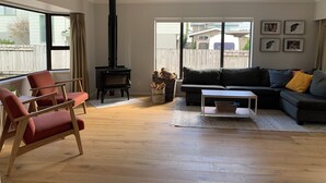 A living room with a sofa, a chair, a fireplace, and a view through a large window. - Relaxing haven for Families - minutes walk to the beach, shops & dining (Tauranga)