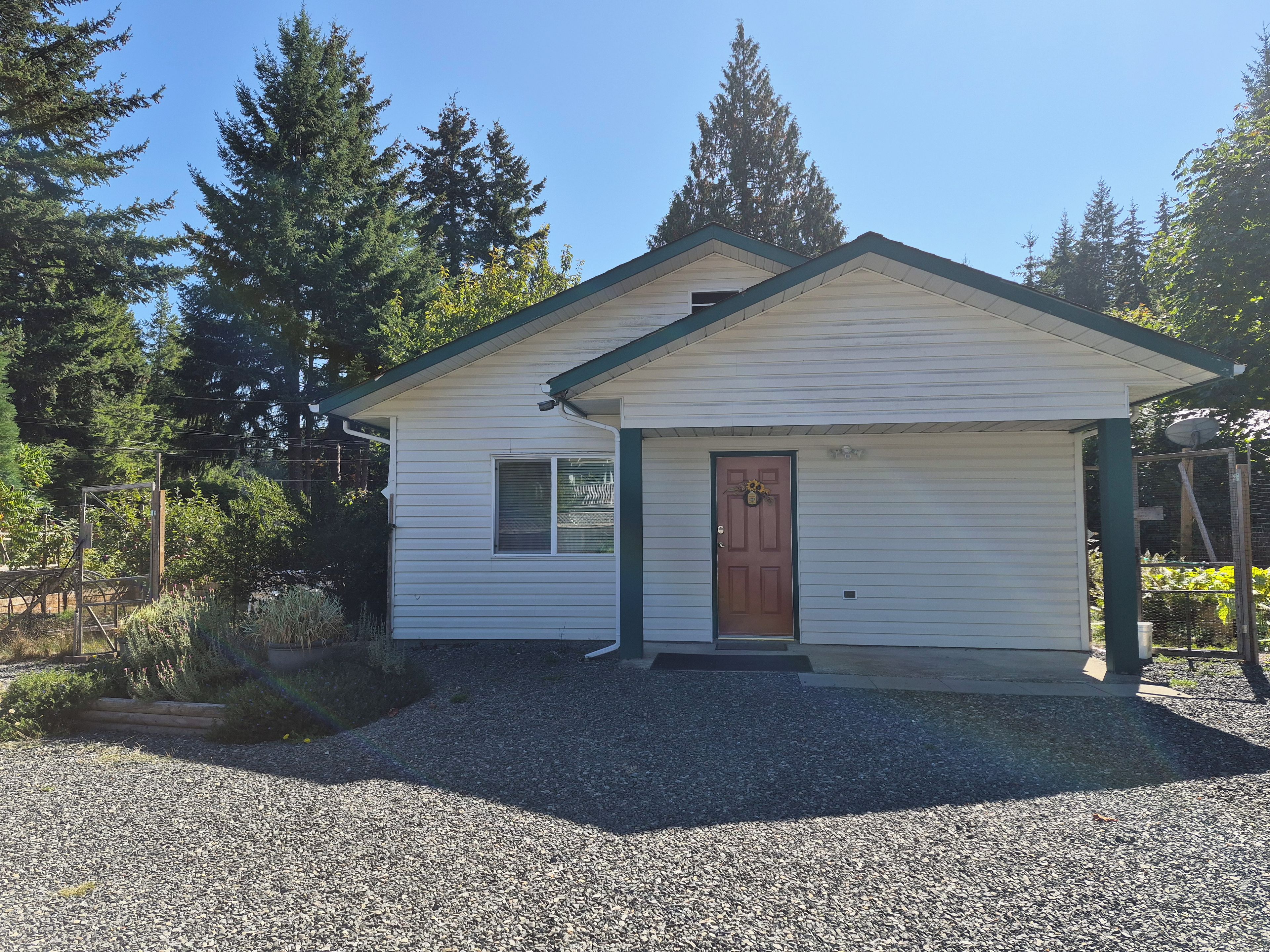 A small, single-story building with a brown door and a window, surrounded by gravel and trees.